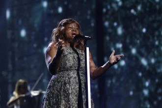 A woman stands at a microphone and sings, her hands outstretched.