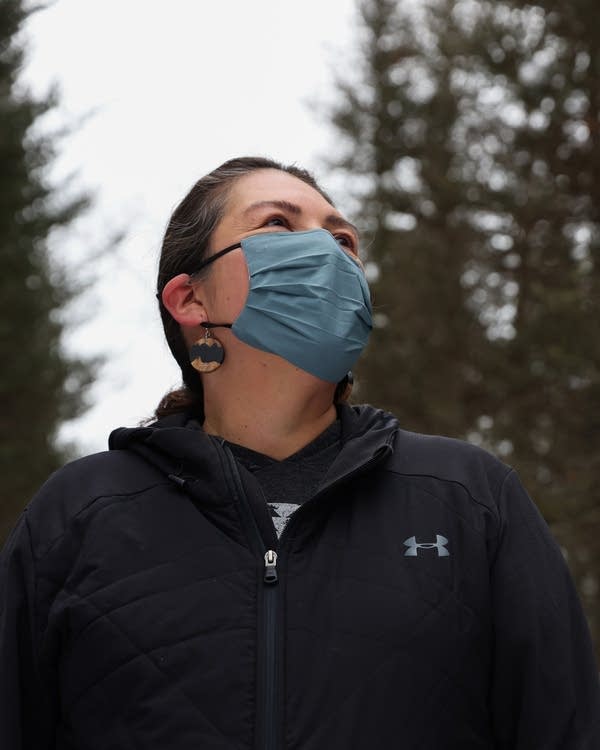 A person wearing a face mask stands outside near pine trees.