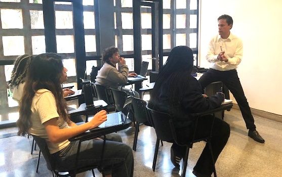 A classroom scene with four people seated at desks and one person standing in front, presenting. Large grid-patterned windows allow natural light into the room.